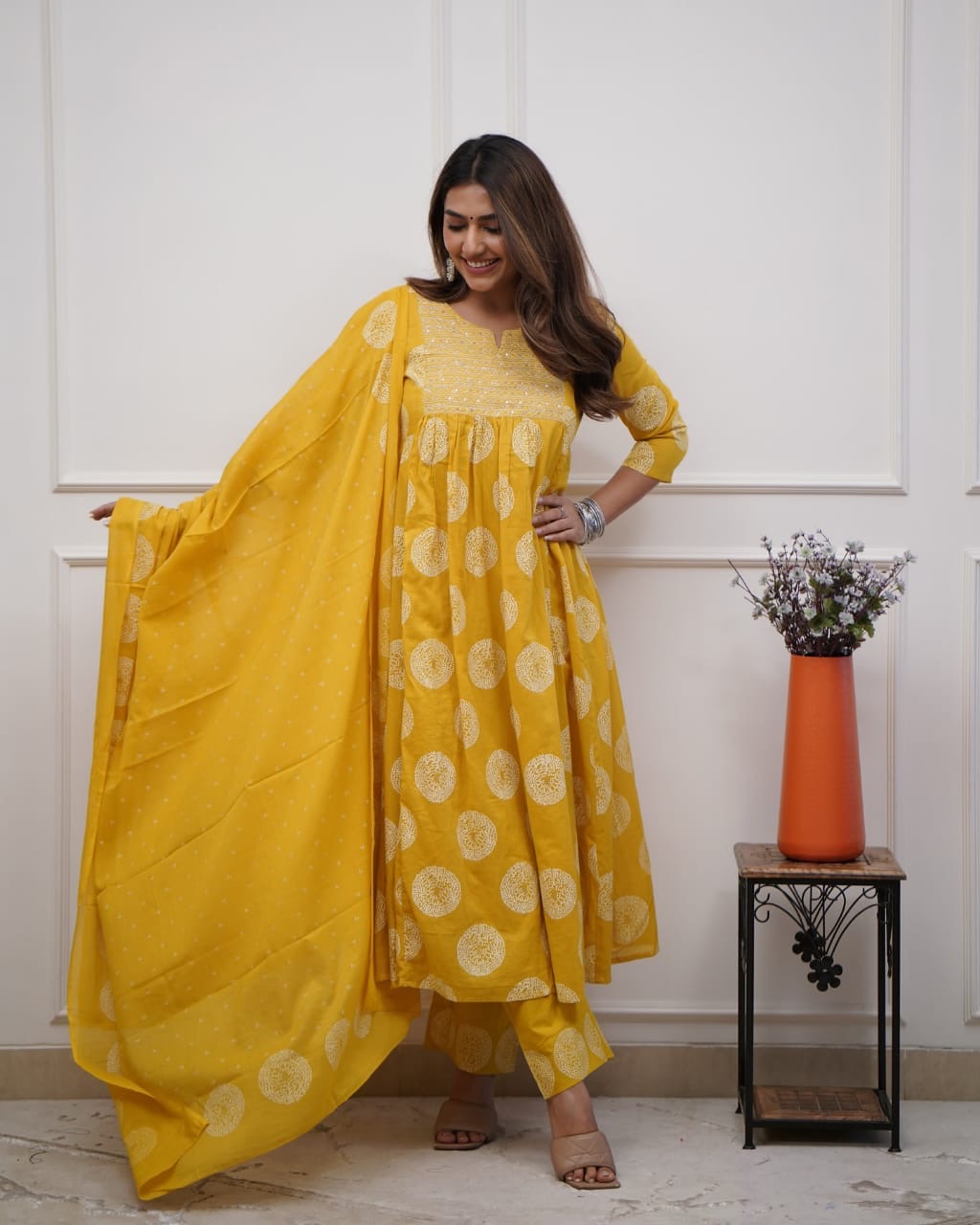Woman wearing a yellow traditional outfit with a white floral pattern, standing indoors.