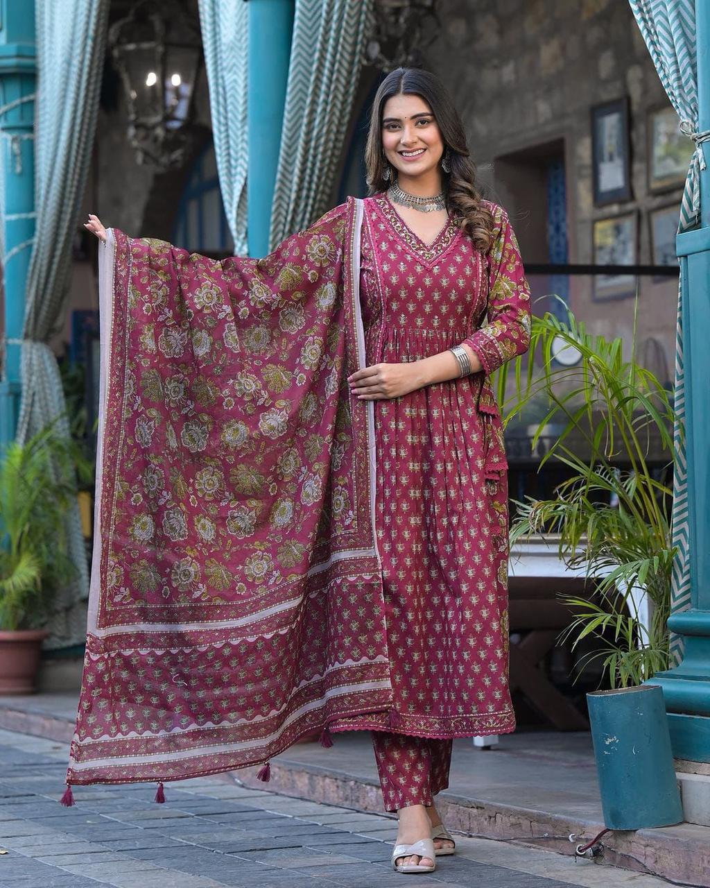 Woman in a traditional maroon and gold outfit holding a matching dupatta, standing in an outdoor setting.