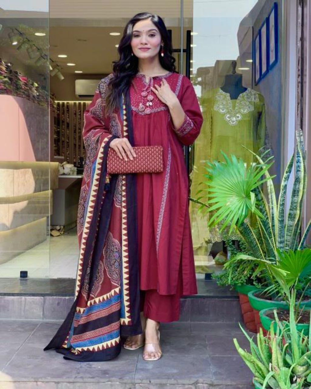 Woman in a red traditional outfit standing in front of a store entrance with plants around.