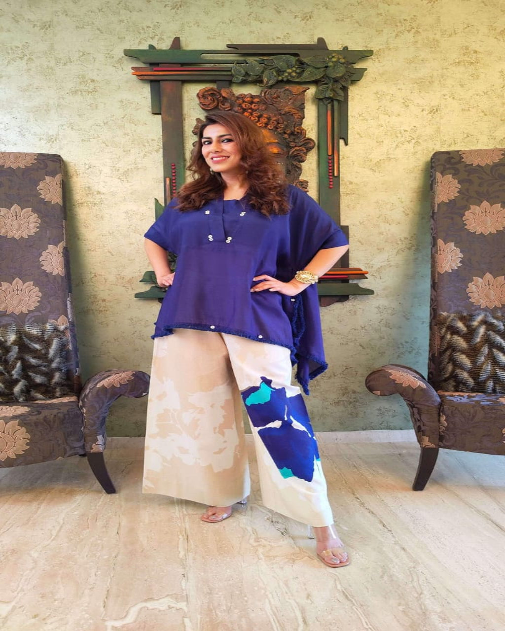 Woman in a blue top and patterned pants standing in a room with decorative chairs and wall art.