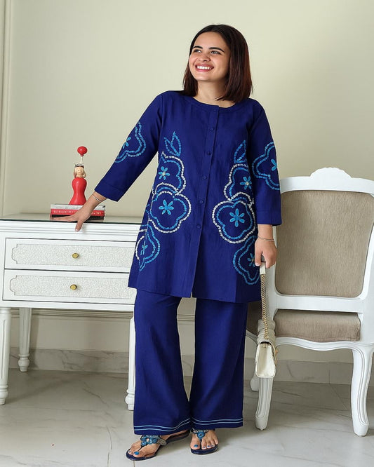 Woman wearing a blue embroidered outfit standing in a room with a white dresser and chair.