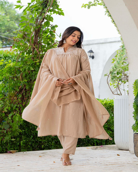 Woman in a beige traditional outfit standing outdoors with greenery in the background