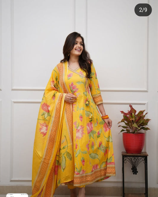 Woman wearing a yellow floral dress with a matching dupatta in a room with a white paneled wall and a plant.