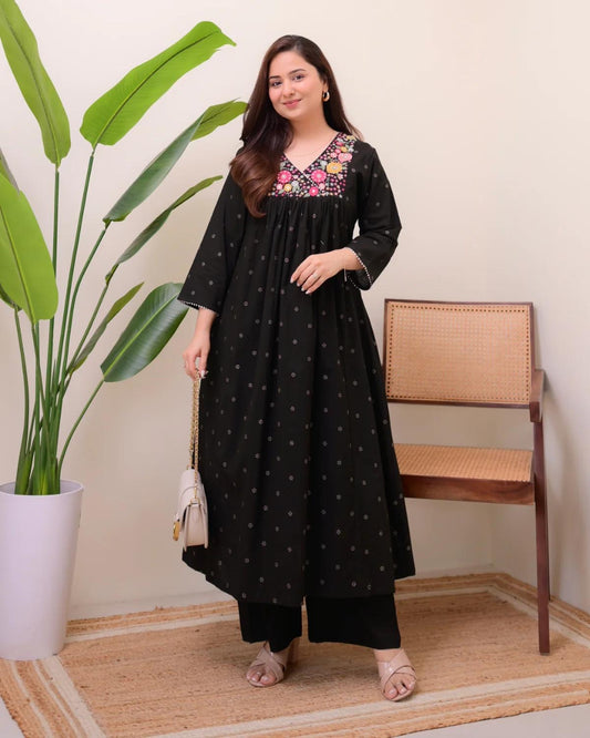 Woman in a black dress with floral embroidery standing next to a plant and chair.