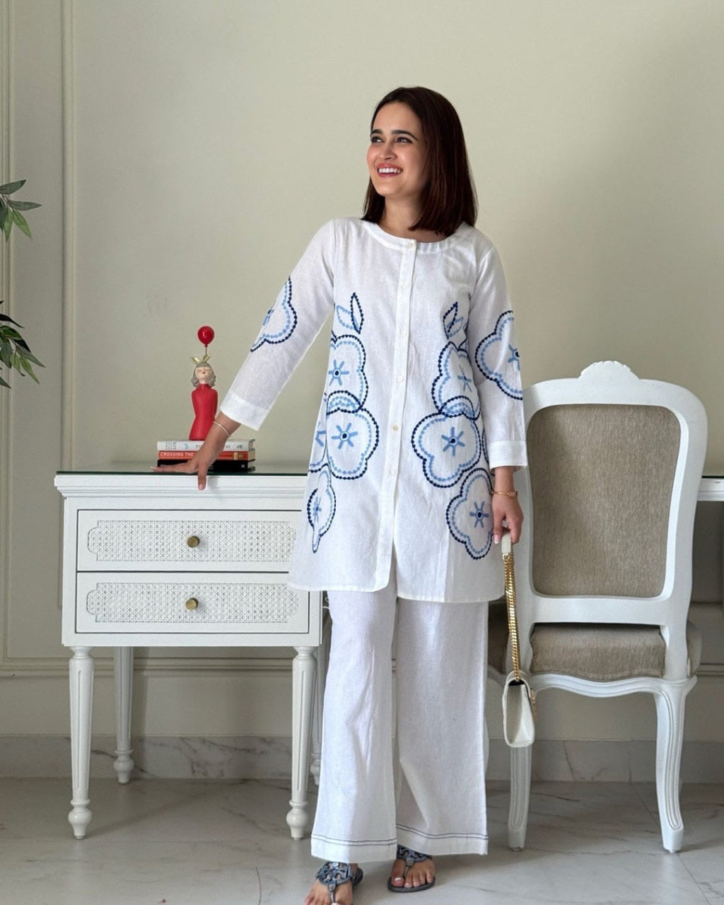 Woman in a white outfit with blue patterns standing in a room with a white chair and table.