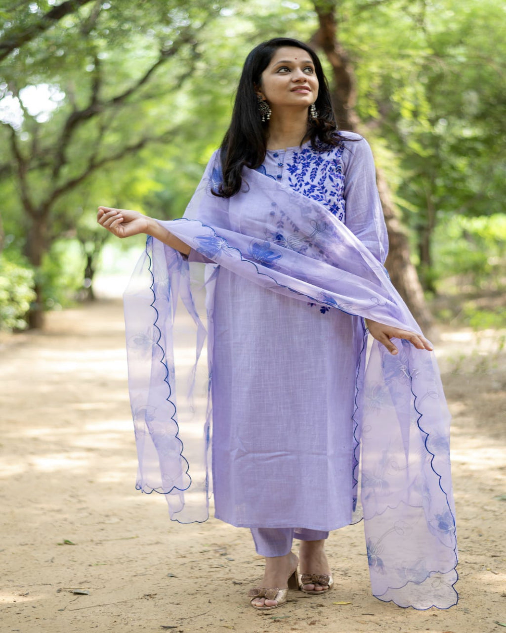 Woman in a light purple traditional outfit with blue patterns standing outdoors.