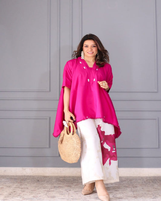 Woman wearing a bright pink top and white pants against a gray wall.